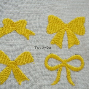 May include: Four yellow embroidered bow designs on a white fabric background. The bows vary in style, with detailed stitching. The text "TodayDo" is visible in the centre of the image. The bows are a bright, cheerful accent.