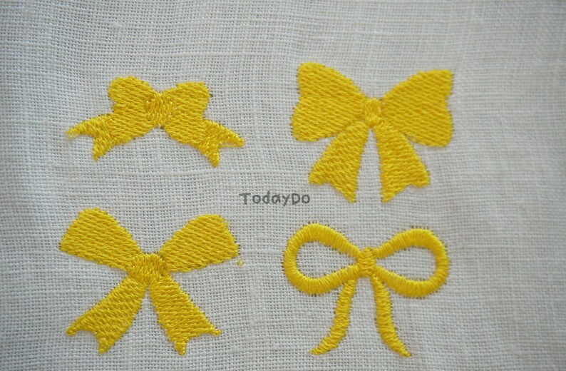 May include: Four yellow embroidered bow designs on a white fabric background. The bows vary in style, with detailed stitching. The text "TodayDo" is visible in the centre of the image. The bows are a bright, cheerful accent.