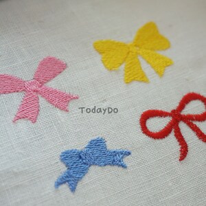 May include: Four embroidered bows in pink, yellow, red, and blue on a white fabric. The bows are detailed with visible stitching, and the text "TodayDo" is visible.