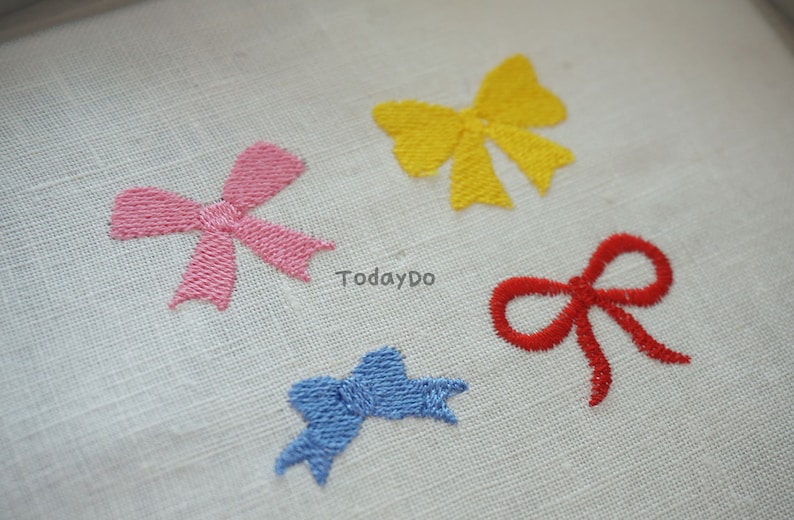 May include: Four embroidered bows in pink, yellow, red, and blue on a white fabric. The bows are detailed with visible stitching, and the text "TodayDo" is visible.