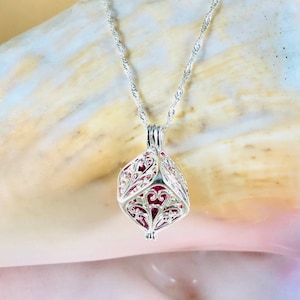 May include: A silver necklace with a filigree pendant. The pendant is a cube shape with intricate cut-out designs and a red interior. The necklace chain is a delicate, twisted style. The background is a seashell.