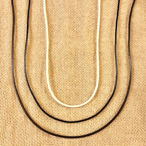 Pu&ograve; includere: Tre cordini di diversi spessori, uno bianco, uno nero e uno marrone, sono disposti su uno sfondo di tessuto beige.
