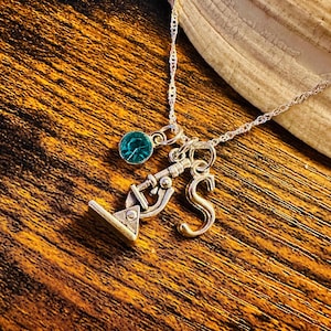 May include: Silver-toned necklace with a microscope charm, a teal-colored gemstone, and a gold-colored letter "S" pendant. The necklace is displayed on a wooden surface, showcasing the jewelry's details.