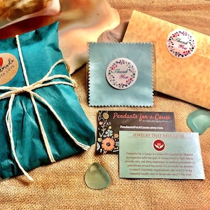 Pu&ograve; includere: Un sacchetto regalo blu turchese legato con uno spago e un'etichetta di carta marrone che dice "fatto a mano con amore". Il sacchetto &egrave; su una superficie di juta con un piccolo ciondolo di vetro blu, un biglietto di ringraziamento blu e bianco, una busta marrone con un adesivo di ringraziamento e un biglietto da visita che dice "Pendants for a Cause" e "Gioielli che salvano vite".
