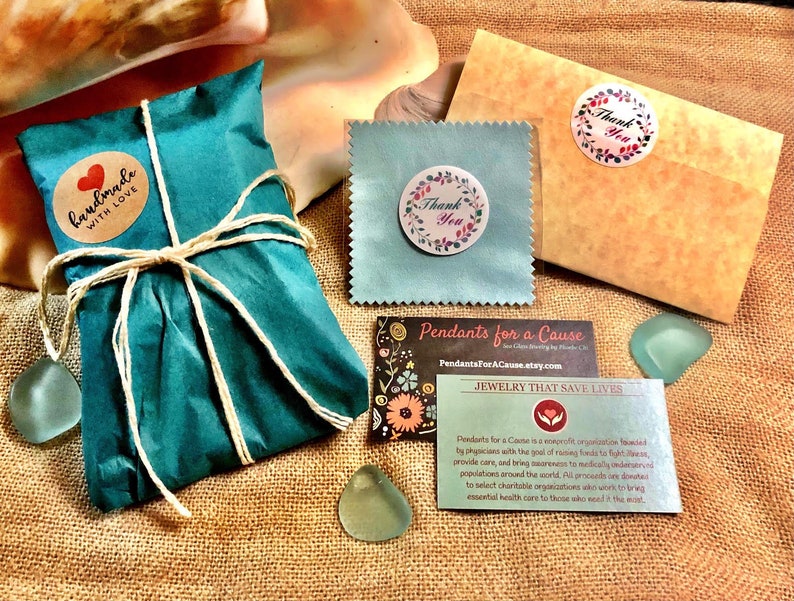 Pu&ograve; includere: Un sacchetto regalo blu turchese legato con uno spago e un'etichetta di carta marrone che dice "fatto a mano con amore". Il sacchetto &egrave; su una superficie di juta con un piccolo ciondolo di vetro blu, un biglietto di ringraziamento blu e bianco, una busta marrone con un adesivo di ringraziamento e un biglietto da visita che dice "Pendants for a Cause" e "Gioielli che salvano vite".