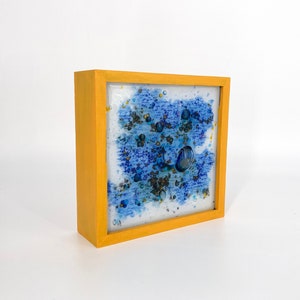 Puede incluir: Una pieza de arte abstracto enmarcada con un fondo azul y blanco y detalles dorados. El marco es de color amarillo brillante.