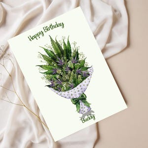 May include: A white card with the text "Happy Birthday" and a green marijuana bouquet wrapped in white and green patterned paper with a green ribbon. The bouquet is tied with a green ribbon and the word "Buddy" is written below the bouquet.