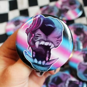 May include: A collection of round buttons with a colorful illustration of a snarling creature's face. The design features shades of pink, blue, and purple against a black background. The buttons are displayed on a black and white checkered fabric.