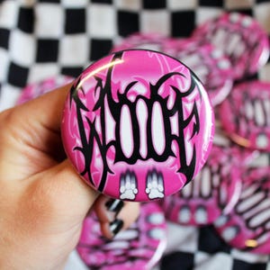 May include: A pink button with the word "WOOF" in black, stylized lettering. The button also features two white paw prints at the bottom. Several similar buttons are visible in the background, set against a black and white checkered fabric.
