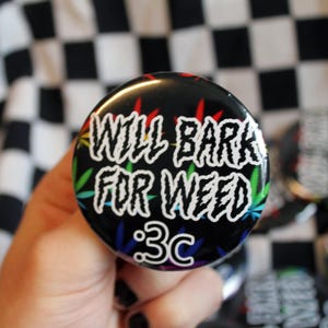 May include: A black button featuring the white text "WILL BARK FOR WEED", with colourful cannabis leaf designs. The button also has the text ":3C" in white. The background is a black and white chequered pattern.