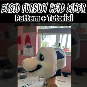 Può includere: Una testa di fursuit in schiuma bianca con una fodera in tessuto nero. La testa è montata su una base in schiuma marrone. La testa è parzialmente assemblata e ha una fodera in tessuto nero. La testa è in fase di lavorazione con forbici e nastro adesivo nelle vicinanze.