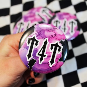 May include: A set of round buttons with a black background and a purple and pink illustration of two figures embracing. The buttons have the letters "T4T" in a gothic font. The background is a black and white checkered pattern.
