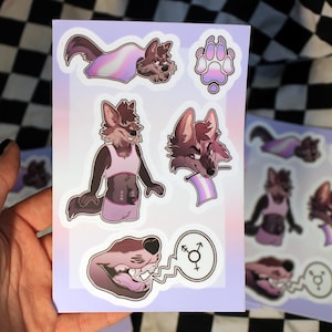 Puede incluir: Una hoja de pegatinas con personajes de lobos antropomórficos en varias poses. Las pegatinas muestran a los personajes de lobos con una bandera transgénero, huellas de patas y bocadillos con símbolos de género. La paleta de colores incluye tonos de marrón, rosa y morado.