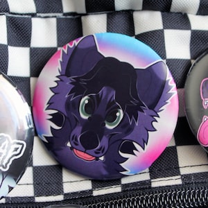May include: A round button featuring a cartoon wolf with purple fur, green eyes, and a pink tongue. The button has a gradient background of pink, blue, and white. Other buttons are visible in the background, set against a black and white checkered pattern.