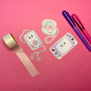 Può includere: Un set di quattro adesivi color pastello con personaggi dei cartoni animati con facce carine. Gli adesivi sono su uno sfondo rosa. I personaggi includono un lettore musicale portatile, una console per videogiochi, un paio di cuffie e un compatto.