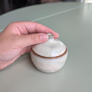May include: A small, round ceramic lidded jar with a glossy off-white glaze and visible brown clay edges. The container features a smooth, curved body and a matching lid with a small, centered knob handle, showcasing a handcrafted aesthetic.