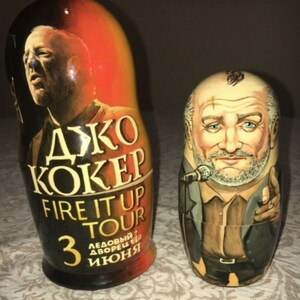 Könnte beinhalten: Zwei hölzerne Nistpuppen, auch Matrjoschka-Puppen genannt, mit einem Porträt des Musikers Joe Cocker. Die größere Puppe ist schwarz und rot bemalt mit dem Text "ДЖО КОКЕР FIRE IT UP TOUR 3 ЛЕДОВЫЙ ДВОРЕЦ ИКОНЫ". Die kleinere Puppe ist braun und beige bemalt mit einem Porträt von Joe Cocker.