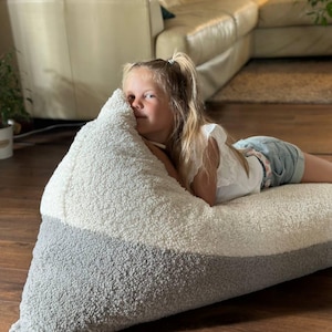 以下が含まれることがあります： ツートンカラーのデザインの、大きくて三角形のビーンバッグチェア。上部は白、下部はグレーです。椅子は柔らかく、質感のある素材で作られています。人がビーンバッグに横たわっています。