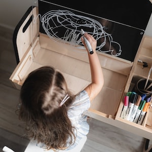 May include: A child draws on a clear dry-erase board attached to a wooden desk. The desk has a built-in storage compartment with various art supplies, including markers, pencils, and paintbrushes.