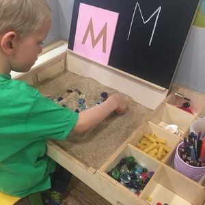 May include: A child plays with a sand table, using colorful stones to spell out the letter "M". The table has a chalkboard with the letter "M" written on it, and a variety of other toys and materials in compartments.