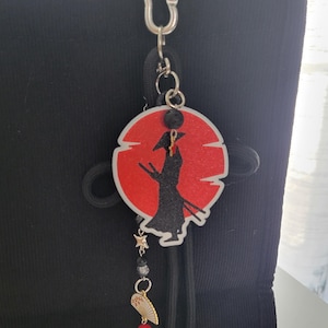 May include: A keychain featuring a circular red glitter background with a black silhouette of a samurai figure. The charm hangs from a silver carabiner and includes a small dangling star, a black bead, and a miniature white and gold fan charm with a red bead.