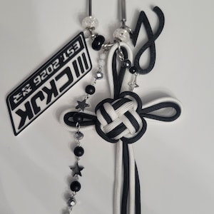 May include: A black and white keychain featuring a knotted cord design, a silver-tone metal clip, and a rectangular tag with the text "MCKJK EST. 2026". The accessory includes a beaded chain with small star-shaped charms and a dangling black letter J.