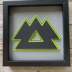 May include: A framed black, green, and yellow paper art piece with a geometric design. The design is a series of overlapping triangles.
