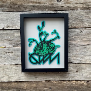 May include: A framed black and green paper art piece with a brain with green vines growing out of it. The brain is sitting on top of the letters "DMN".