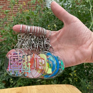 May include: A collection of clear acrylic keychains with colorful, abstract designs. Each keychain has a silver metal ring and clasp. The designs feature a variety of colors, including pink, blue, green, and yellow. The keychains are held in a hand.