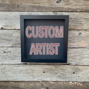 May include: A black framed sign with the words "CUSTOM ARTIST" in pink outlined letters on a black background.