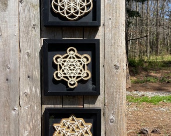 May include: Three framed geometric art pieces. Each piece features a layered design in natural wood tones set against a black background. The top frame displays a Flower of Life pattern, the middle a Metatron's Cube, and the bottom a Star Tetrahedron.