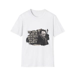 May include: A white short-sleeved t-shirt featuring a black and white graphic of Albert Camus with a cigarette. The text reads: YOU THINK LIFE IS BEAUTIFUL BECAUSE YOU DON'T KNOW IT. A street lamp and dark ink-like background elements are visible.