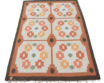 May include: A vintage rug with a geometric pattern of orange, brown, and white flowers. The rug has a brown border and a fringe.