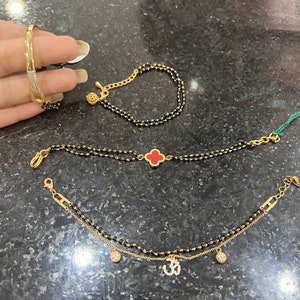 May include: Three gold bracelets with black beads. The top bracelet has a small gold ball charm. The middle bracelet has a red clover charm. The bottom bracelet has two small gold ball charms and an Om symbol charm.