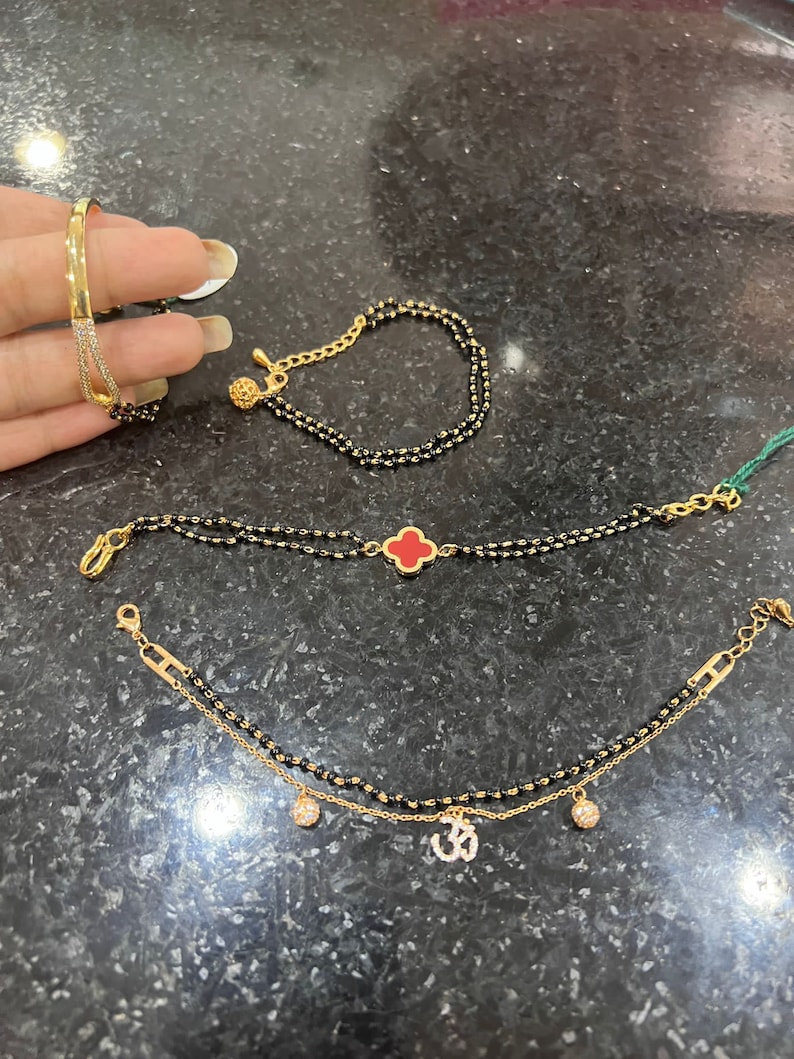 May include: Three gold bracelets with black beads. The top bracelet has a small gold ball charm. The middle bracelet has a red clover charm. The bottom bracelet has two small gold ball charms and an Om symbol charm.
