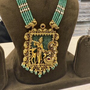 May include: A gold-toned necklace with a large pendant featuring a figure playing a flute, a deer, and a peacock. The pendant is framed with intricate designs and adorned with emerald green beads and red accents. The necklace has strands of green beads and white pearls.