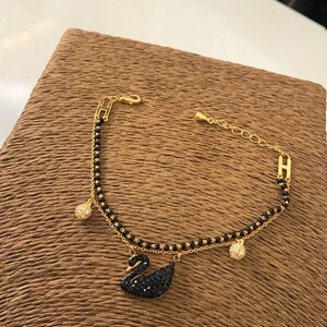 May include: A gold chain bracelet with a black swan charm and two black bead charms. The swan charm is covered in black crystals.