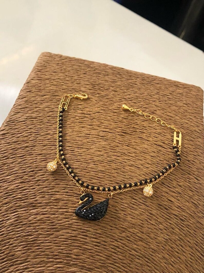 May include: A gold chain bracelet with a black swan charm and two black bead charms. The swan charm is covered in black crystals.