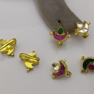 Puede incluir: Cinco amuletos de metal dorado con piedras de diferentes colores y diseños. Cada amuleto tiene forma de media luna con un pequeño lazo en la parte superior. Las piedras son rosas, verdes y blancas.