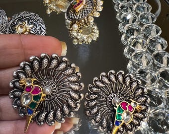 May include: A pair of silver and gold earrings with a peacock design. The earrings feature a colourful bird with green, blue, pink, and white stones.