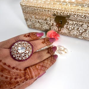 May include: A large, ornate ring with a large, clear gemstone surrounded by smaller, clear gemstones. The ring is set in a gold-tone metal and has a band of small, red gemstones.