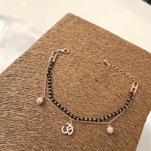 May include: A delicate gold bracelet with a black beaded chain and two charms. One charm is a small, round, clear crystal, and the other is a larger charm with a diamond-encrusted Om symbol.