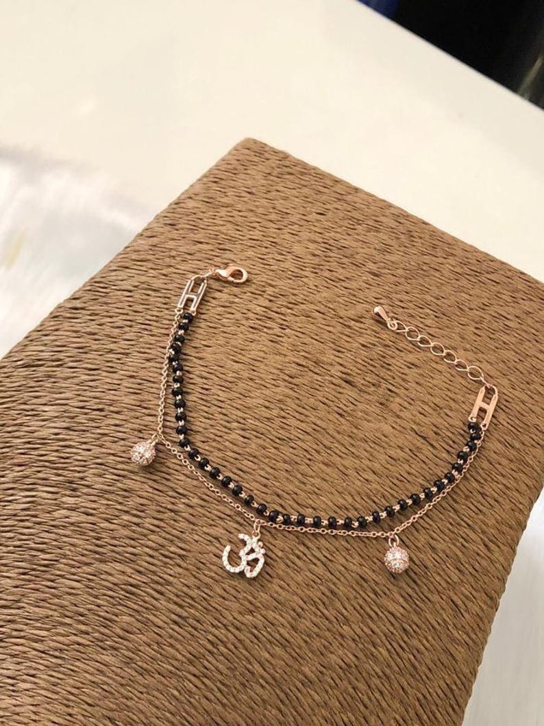 May include: A delicate gold bracelet with a black beaded chain and two charms. One charm is a small, round, clear crystal, and the other is a larger charm with a diamond-encrusted Om symbol.