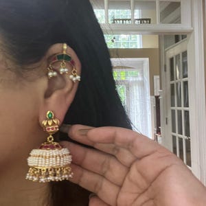 Może przedstawiać: Złote kolczyki w stylu jhumka, z czerwonymi i zielonymi akcentami oraz perłową frędzlą. Widoczny jest również pasujący nausznik z zielonymi, czerwonymi i białymi detalami. Biżuteria jest noszona na uchu osoby.