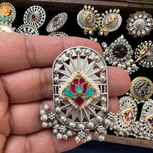 May include: A silver and white metal earring with a floral design and a turquoise and pink stone in the center. The earring has a pearl drop and is surrounded by a decorative border.