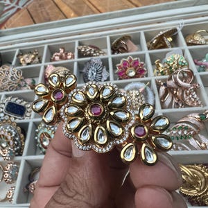 May include: A gold-toned floral ring with clear and red gemstones. The ring is held in front of a jewelry display case filled with various rings. The ring has a central flower design with smaller flowers on either side.