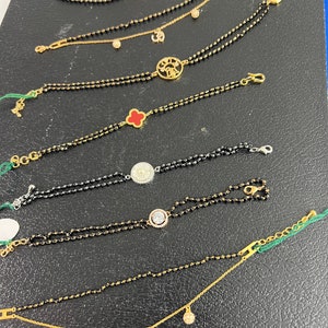 May include: A collection of nine gold bracelets with black beads and various charms, including a clover, a circle with a pearl, a circle with diamonds, a swan, a sun, and an Om symbol.