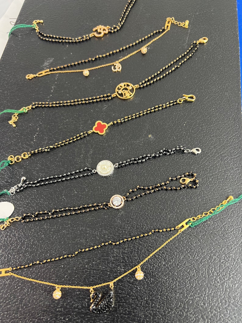May include: A collection of nine gold bracelets with black beads and various charms, including a clover, a circle with a pearl, a circle with diamonds, a swan, a sun, and an Om symbol.
