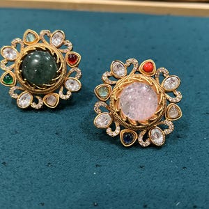 May include: Two gold-toned rings with intricate floral designs and colorful gemstones. The ring on the left has a green gemstone, while the ring on the right has a pink gemstone.