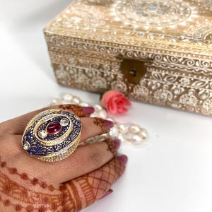 May include: A gold ring with a blue and white enamel design and red gemstones. The ring is oval shaped and has intricate details.